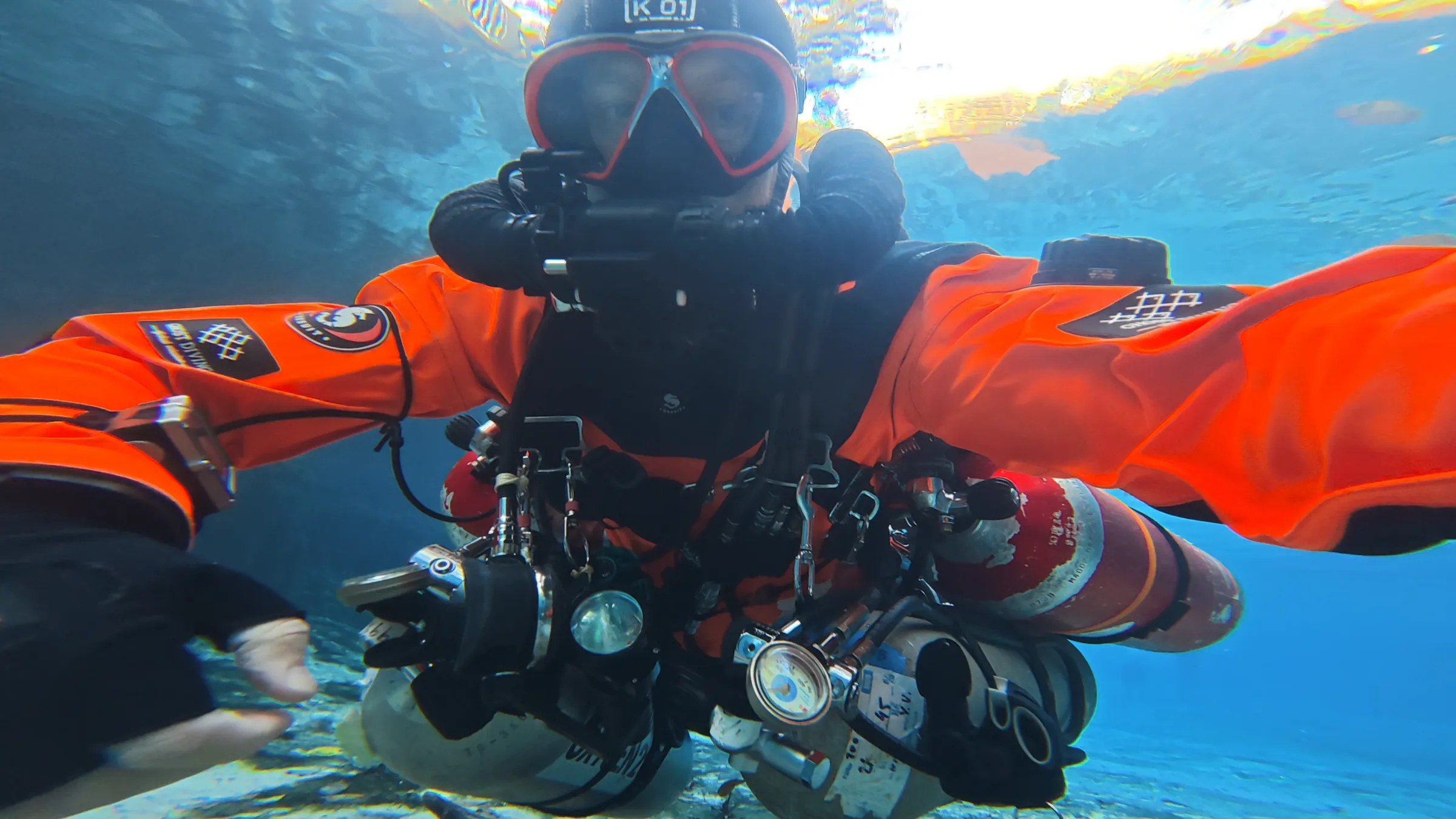 A diver in an orange drysuit reaches toward the camera in sunlit turquoise water